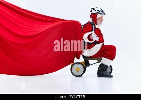 Der Weihnachtsmann auf dem Dreirad des Kindes hält eine riesige, schwere rote Tasche voller Geschenke hinter sich, mit fröhlichem Ausdruck vor weißem Atelierhintergrund. Stockfoto