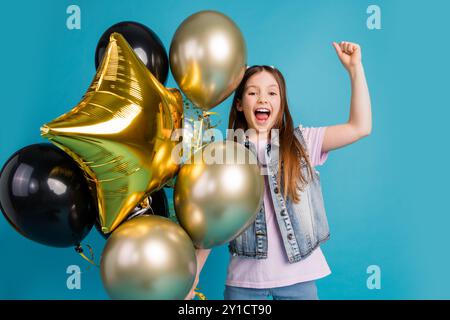 Foto-Porträt des entzückenden kleinen Mädchens halten Luftballons gewinnen gekleidete stilvolle Denim Kleid isoliert auf blauem Farbhintergrund Stockfoto