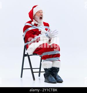 Santa-Duell am Stuhl, mit breiten Augen und schockiert, verzweifelt versucht, sich von silbernem Klebeband zu befreien, das ihn an Ort und Stelle hält. Stockfoto