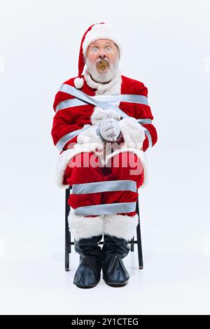 Der Weihnachtsmann ist fest in Klebeband gewickelt, mit Tannenzapfen in den Mund gesteckt, seine Augen weit unglauben, während er unbeweglich auf dem Stuhl sitzt. Stockfoto