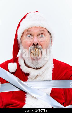 Mit Tannenzapfen im Mund sitzt der Weihnachtsmann in Klebeband gebunden, sieht verwirrt und regungslos aus, seine Hände und Füße sind sicher am Stuhl befestigt. Stockfoto