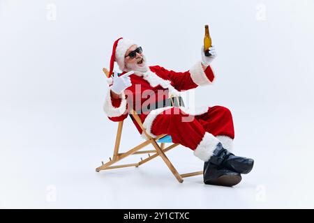 Der Weihnachtsmann loungt im Stuhl, Sonnenbrille an, hebt die Bierflasche auf, lächelt selbstbewusst, während er stolz auf die Flasche vor weißem Hintergrund zeigt. Stockfoto
