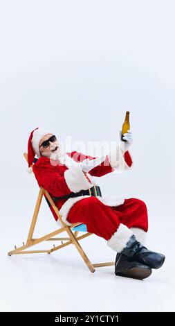 Der Weihnachtsmann lehnt sich zurück im Stuhl, genießt Bier in der Hand, trägt eine Sonnenbrille und sieht entspannt und sorglos vor weißem Hintergrund aus. Stockfoto