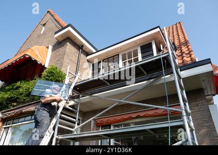 Der Bauarbeiter steigt eine Leiter mit einem neuen Fenster mit Stangenteilung hinauf. Renoviertes Haus mit Doppelverglasung Stockfoto