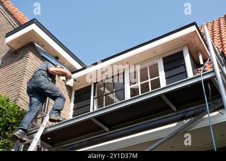 Der Bauarbeiter steigt eine Leiter mit einem neuen Fenster mit Stangenteilung hinauf. Renoviertes Haus mit Doppelverglasung Stockfoto