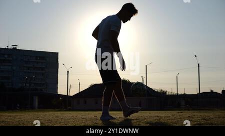 Profifußballer jongliert Fußball im Stadion bei Sonnenuntergang. Junger Mann, der auf dem grünen Feld Ball tritt. Sportler, der mit ihm Tricks auf der Wiese übt Stockfoto