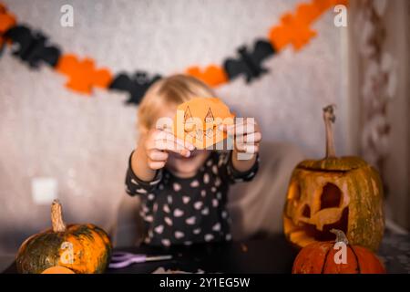 Ein kleines Kind zeigt einen leuchtenden orangefarbenen Kürbisausschnitt, umgeben von geschnitzten Kürbissen und festlichen Dekorationen. Kunstzubehör und Halloween-Kunsthandwerk Stockfoto