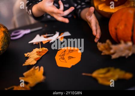 Ein Kind bastelt mit Begeisterung Halloween-Dekorationen aus orangefarbenem Papier, umgeben von Herbstblättern und Kürbissen. Die Atmosphäre spiegelt ein gemütliches cr wider Stockfoto