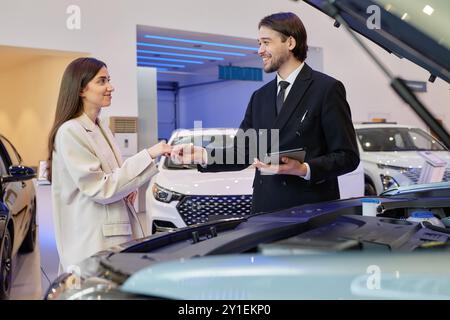 Seitenansicht eines lächelnden zentralasiatischen Autohändlers, der weibliche Kunden Hand nimmt und einen Deal abschließt, während glückliche Frau im Händlerzentrum ein neues Auto kauft Stockfoto