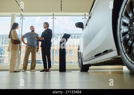 Bild vom Boden aus, in dem ein lächelnder Autohändler Kunden über neue Premium-Fahrzeuge berät und die Einkaufsbedingungen mit einem Paar im Ausstellungsraum neben einem riesigen Fenster im Händlerzentrum, Kopierraum, bespricht Stockfoto