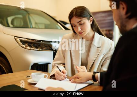 Mittlere Aufnahme der jungen modischen Frau, die einen Kaufvertrag am Schreibtisch unterzeichnet und ein neues Auto mit Hilfe des Autohändlers im Händlerzentrum kauft, Kopierraum Stockfoto