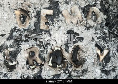 Verdorbene Holzbriefe, in denen das Wort Demokratie steht, sitzen unter einem Aschenstapel in einem Mülleimer und deuten darauf hin, dass die Demokratie angegriffen wird. Stockfoto