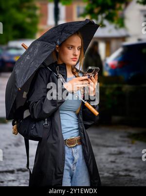 Eine junge Frau steht im Regen unter einem schwarzen Regenschirm und konzentriert sich auf ihr Smartphone, während sie einen schwarzen Regenmantel und eine blaue Jeans trägt. Stockfoto
