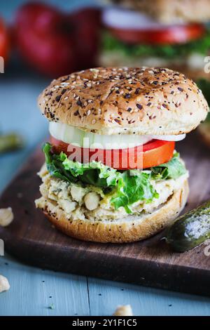 Ein veganes vegetarisches Sandwich mit Kichererbsenaufstrich, Salat, Scheiben von frischen Tomaten und Zwiebeln mit Gurken auf der Seite über allem Bagel Brötchen Stockfoto