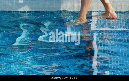 Beine und Füße Eines Mannes, der über Steps In Einen blau gefliesten Swimmingpool einsteigt Stockfoto