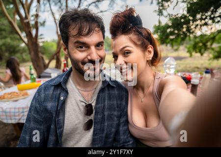 Nahaufnahme eines fröhlichen Paares, das ein Selfie bei einem Picknick im Freien macht. Junger Mann und Frau, die vor der Kamera lächeln, mit Picknicktisch und Natur im Hintergrund, c Stockfoto