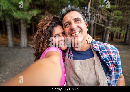 Fröhliches Paar mittleren Alters, das ein Selfie in einem Kiefernwald macht. Lächelnde Männer und Frauen, die sich umarmen, Glück und Liebe in Reifen Beziehungen zeigen. E Stockfoto