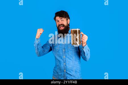 Glücklicher Mann mit Bierkrug, der den Sieg mit erhobener Hand feiert. Bärtiger Mann in Denim-Hemd, der leckeres Bier vom Fass in Pub oder Bar trinkt. Gutaussehender männlicher wi Stockfoto