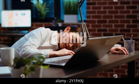Müder Angestellter, der auf dem Schreibtisch geschlafen hatte, nachdem er die ganze Nacht mit dem Schreiben von E-Mails zu kämpfen hatte. Schläfrige Geschäftsfrau, die im Büro schläft, nachdem sie über Nacht gearbeitet hat, im Laptop geschlafen hat, Kamera B Stockfoto