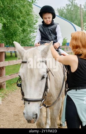 Kind in schwarzem Schutzhelm und Weste, das auf dem Bauernhof auf einem weißen Pferd reitet, Frau mit Zügen, während sie das Kind auf einem umzäunten Feldweg führt, grüne Bäume im Hintergrund Stockfoto