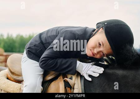 Junge in Reitkleidung, während er mit einem Lächeln auf einem Pferd sitzt, im Freien mit grünen Bäumen im Hintergrund, die Interaktion zwischen Kind und Tier während der Reittätigkeit zeigen Stockfoto