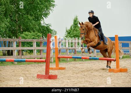 Reiter, der das Pferd über gemalte Hindernisse in einer Reitarena im Freien mit üppig grünen Bäumen im Hintergrund und Sandboden führt, zeigt Reitfertigkeiten Stockfoto