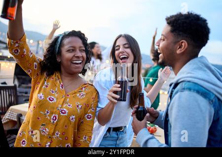 Gruppe von drei verschiedenen jungen, aufgeregten Freunden, die zusammen tanzen und Bier auf einer Sommerparty auf dem Dach halten Stockfoto