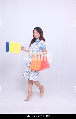 Ganzkörper Junge indonesische Frau lacht in einem modernen chinesischen Kleid, während sie beim Einkaufen Papiertüten in vielen Farben auf einem trägt Stockfoto