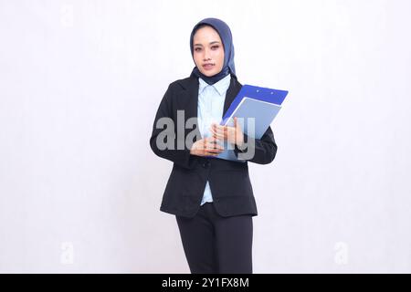 Exklusive indonesische Bürofrau mit Hijab steht elegant mit Stift, Klemmbrett und Buch. Schöne muslimische Frau in blauem Hemd Stockfoto