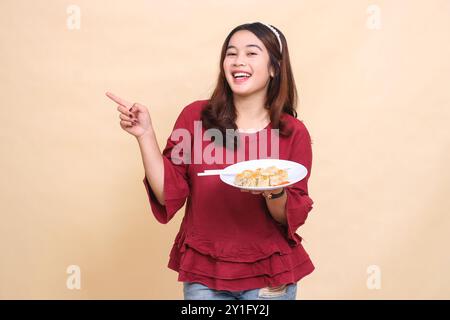 Die schöne junge indonesienfrau in rotem Hemd ist glücklich, einen Teller mit Dimsum (chinesisches Essen) und Essstäbchen zu tragen, während sie nach rechts zeigt. US Stockfoto