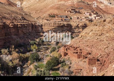 Blick auf ein Dorf mit steilem Schotterweg in das trockene, trockene Tal mit karger Vegetation jenseits traditioneller berber-Lehm-Steinhäuser, zerklüftete Felswände b Stockfoto