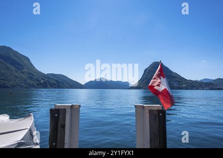 Lugano. Schweiz. Kanton Tessin. Blauer Himmel an einem sonnigen Tag mit der Schweizer Flagge über dem Luganer See. Blick von einer Holzbrücke Stockfoto