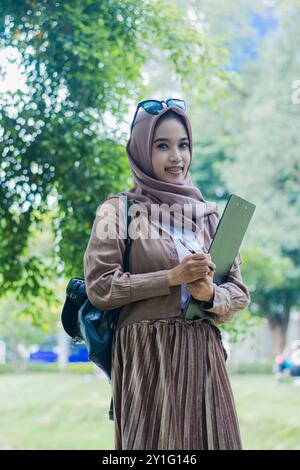 Reife asiatische Frau im Hijab lächelt die Kamera an und trägt das Stiftenbuch vor dem Klemmbrett, die Tasche, die im Park steht. Studentinnen für das Thema Stockfoto