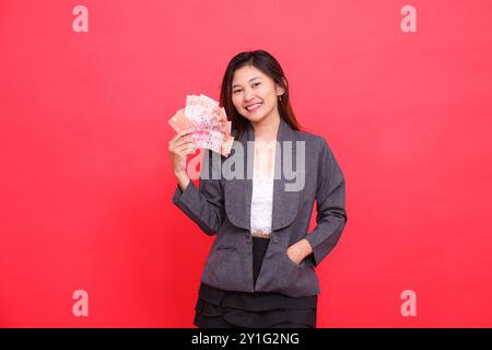 Ausdruck einer fröhlichen indonesischen Bürofrau, die Debitkreditkarte und Geld aufrecht hält und in der Tasche in die Kamera blickt, die graue Jacke und Rot trägt Stockfoto