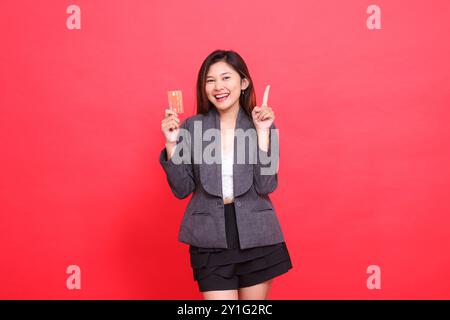 Fröhliche indonesische Bürofrau in die Kamera, die eine Debit-Kreditkarte hält, während sie mit Jacke und Rock auf einer roten Ba nach oben zeigt Stockfoto