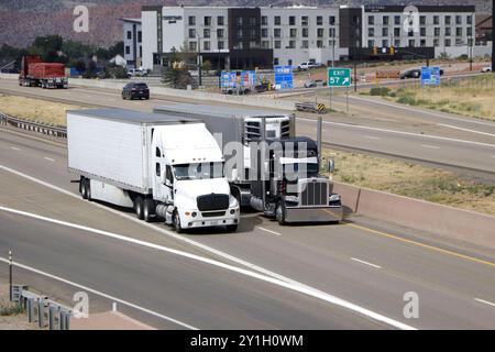Zwei große Sattelzüge fahren nebeneinander auf einer mehrspurigen Autobahn und einem Gebäudekomplex im Hintergrund. Stockfoto