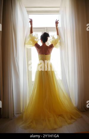 Die Silhouette einer Frau in einem goldenen, luxuriösen Kleid vor dem Hintergrund eines Fensters hält einen Vorhang mit den Händen. Elegante Dame in gelber Seide Stockfoto