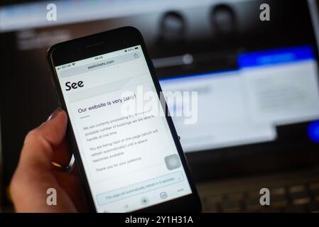 Eine Hand hält einen Handybildschirm mit der Meldung „siehe Tickets Website“ für Tickets für Oasis Reunion. Ticketmaster-Bildschirm auf Laptop im Hintergrund. Stockfoto