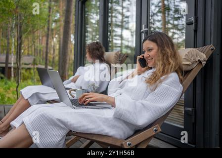 Zwei Frauen in weißen Bademänteln entspannen auf der Terrasse und arbeiten an Laptops. Einer telefoniert und genießt eine ruhige Waldumgebung. Stockfoto