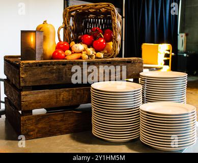 Eine rustikale Ausstellung mit einer Holzkiste gefüllt mit frischem Gemüse wie Tomaten, Kürbis und Pilzen. In der Nähe befindet sich ein Stapel weißer Platten Stockfoto