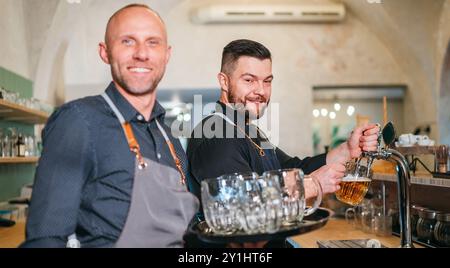 Stilvoller bärtiger Barkeeper in schwarzer Uniform, der Bier an der Bar anzapft und der Kellner mit Tablett plauderte miteinander und lächelte miteinander. Erfolgreiches People Team Wo Stockfoto