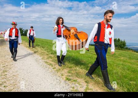 Eine Gruppe von Musikern in traditioneller Kleidung, die auf einem Landweg mit Kontrabass unter blauem Himmel spazieren Stockfoto