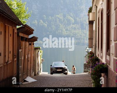 Hallstatt, Österreich - 31. August 2024: Eine ruhige Gasse führt zu einem Seeblick mit einem Auto am Wasserrand, blumengeschmückten Gebäuden und einem Stockfoto