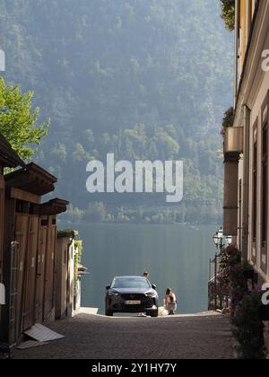 Hallstatt, Österreich - 31. August 2024: Eine ruhige Gasse führt zu einem Seeblick mit einem Auto am Wasserrand, blumengeschmückten Gebäuden und einem Stockfoto