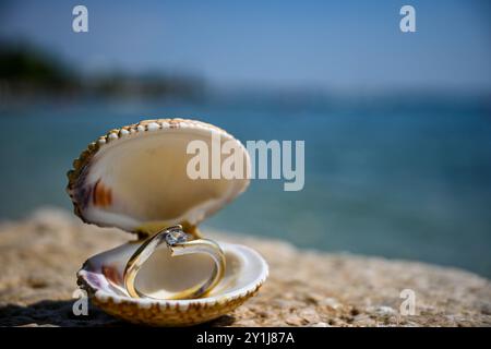 Der wunderschöne Verlobungsdiamantring wird in einer offenen Muschel am Strand mit dem Meer im Hintergrund präsentiert. Stockfoto