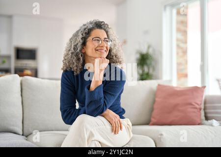 Glückliche 50-jährige Frau, die zu Hause auf dem Sofa sitzt und wegschaut, Porträt. Stockfoto