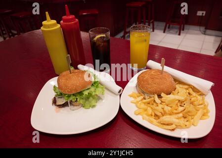 Mahlzeit für zwei in einem Fast Food: Zwei leckere gegrillte Burger mit Pommes frites, Limonade und Orangensaft auf weißen Gerichten und rotem Holztisch, mit Plastikscheiben Stockfoto