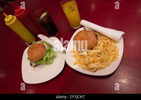 Mahlzeit für zwei in einem Fast Food: Zwei leckere gegrillte Burger mit Pommes frites, Limonade und Orangensaft auf weißen Gerichten und rotem Holztisch, mit Plastikscheiben Stockfoto