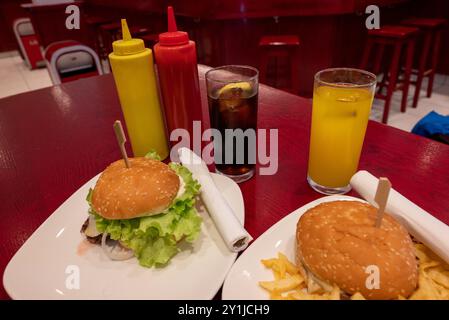 Mahlzeit für zwei in einem Fast Food: Zwei leckere gegrillte Burger mit Pommes frites, Limonade und Orangensaft auf weißen Gerichten und rotem Holztisch, mit Plastikscheiben Stockfoto