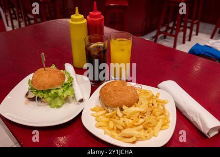 Mahlzeit für zwei in einem Fast Food: Zwei leckere gegrillte Burger mit Pommes frites, Limonade und Orangensaft auf weißen Gerichten und rotem Holztisch, mit Plastikscheiben Stockfoto